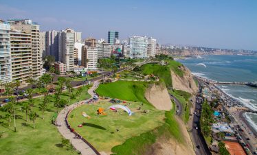 A scenic view of Miraflores, Lima, showing lush green cliffs, modern buildings, and paragliders soaring over the Pacific Ocean.