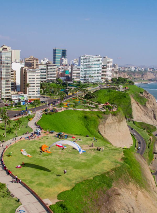 A scenic view of Miraflores, Lima, showing lush green cliffs, modern buildings, and paragliders soaring over the Pacific Ocean.