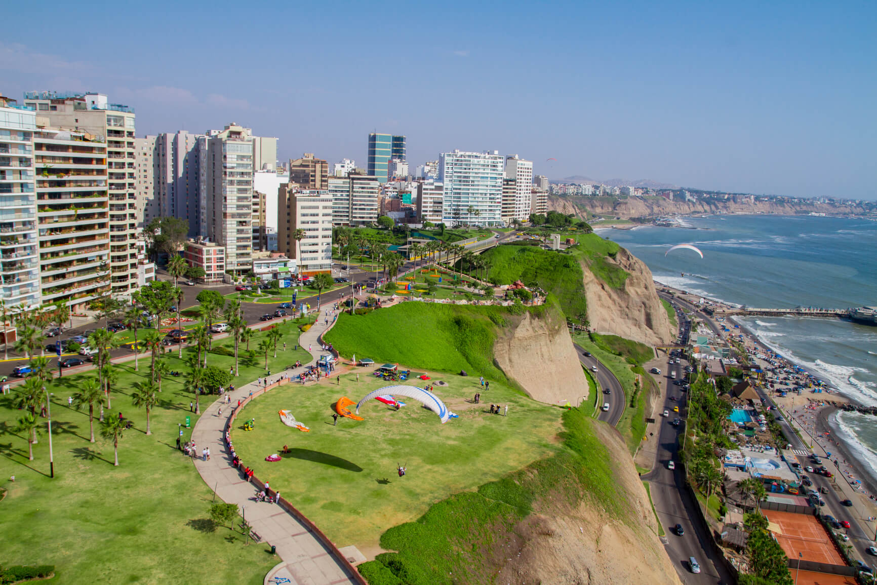 A scenic view of Miraflores, Lima, showing lush green cliffs, modern buildings, and paragliders soaring over the Pacific Ocean.