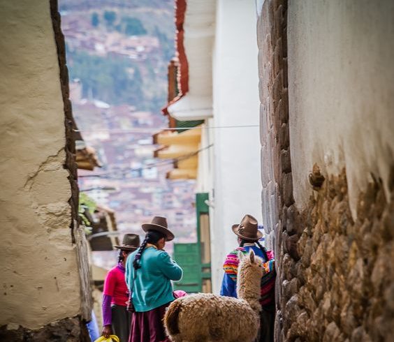 A traditional Andean scene in Cusco, Peru, featuring local women in colorful attire, a llama, and colonial-style architecture with the city’s hills in the background.