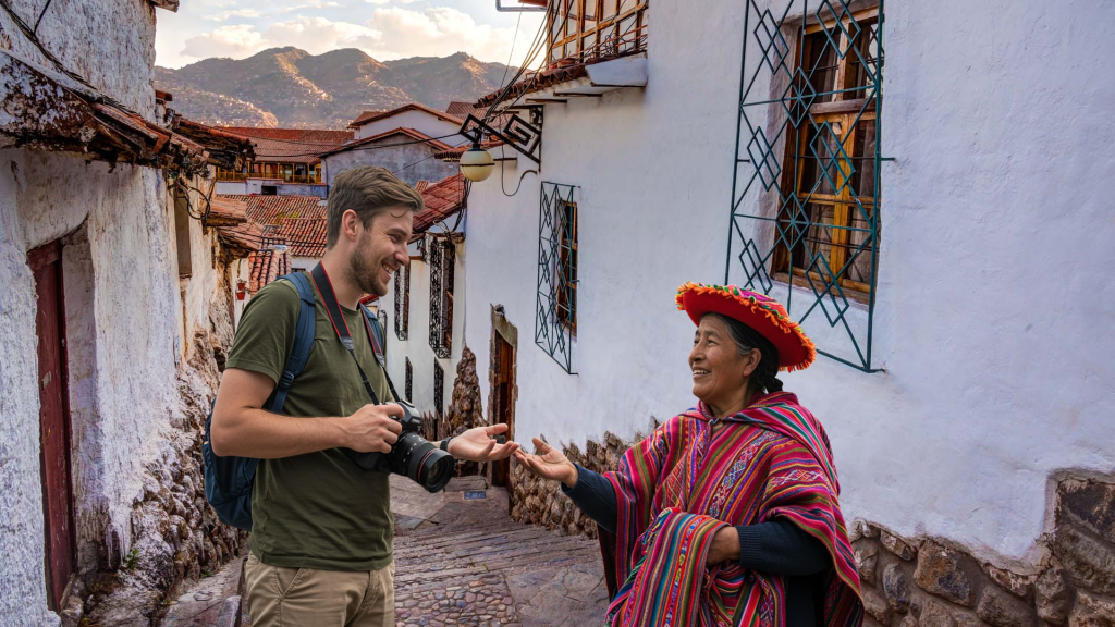A smiling tourist with a backpack and camera interacts with an indigenous Peruvian woman in traditional Andean clothing on a picturesque cobblestone street in Cusco. They exchange a small object while sharing a joyful moment.