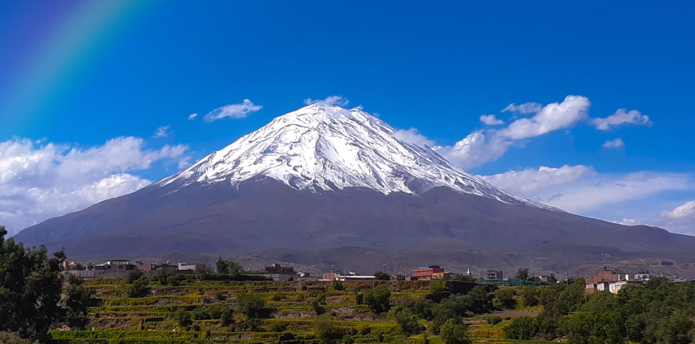 Vista panorámica del majestuoso volcán Misti en Arequipa, Perú, con su cima nevada, cielos despejados y un arcoíris en el cielo. En el primer plano, se observan andenes verdes y cultivos.