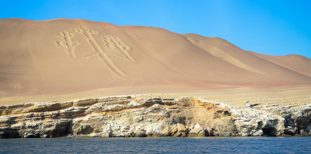 Vista del geoglifo El Candelabro de Paracas, grabado en la arena de una colina costera en la Reserva Nacional de Paracas, Perú. En primer plano, el océano y acantilados rocosos complementan el paisaje desértico.