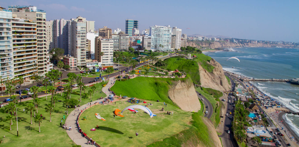 Vista panorámica de los acantilados de Miraflores en Lima, Perú, con parapentes sobrevolando el malecón y el Océano Pacífico, rodeado de áreas verdes y modernos edificios.