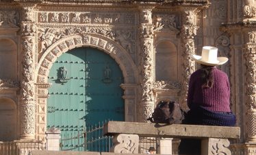 A woman wearing a traditional white hat and purple sweater sits on a stone bench, gazing at the ornate façade of a historic church in Cajamarca, Peru