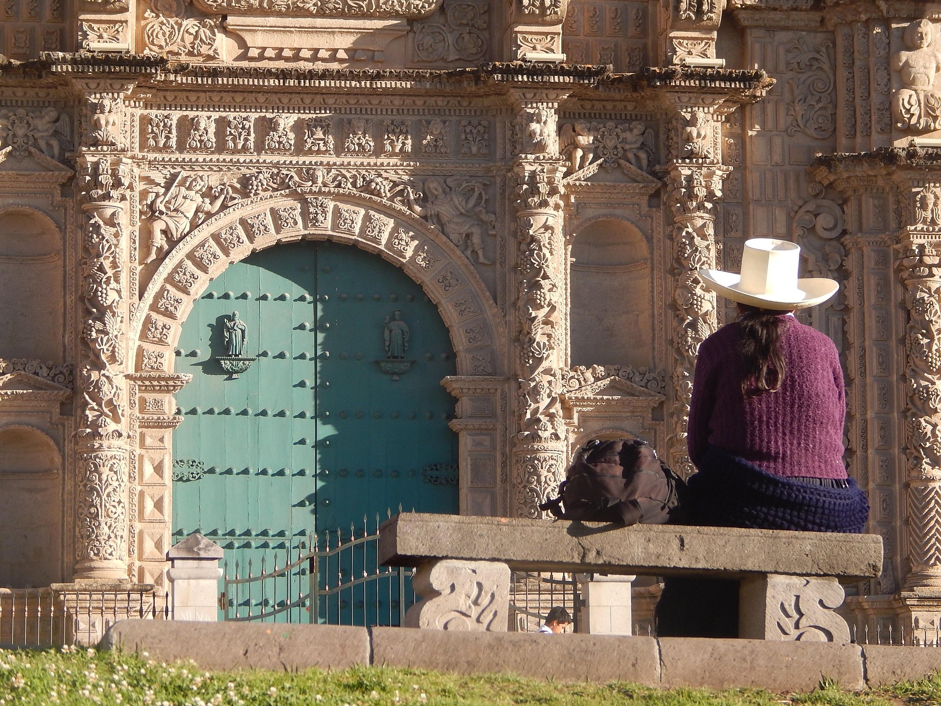 A woman wearing a traditional white hat and purple sweater sits on a stone bench, gazing at the ornate façade of a historic church in Cajamarca, Peru